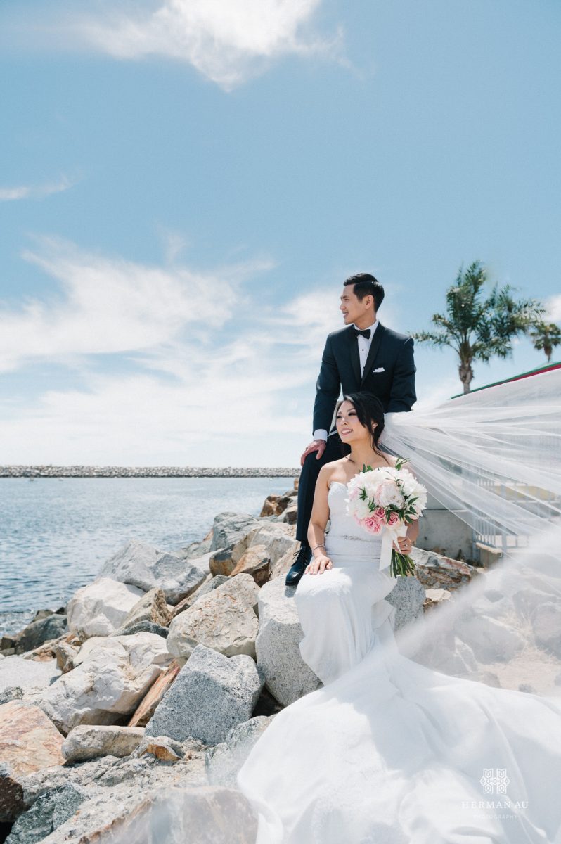 Bride and groom sitting on the rocks with the Redondo Beach Marina in the background and the bride's veil floating in the wind